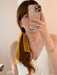 A woman with brown hair in a ponytail, tied with Noémiah’s “Three Ribbons,” takes a mirror selfie. She wears a white top, gold jewelry, and holds her phone with a cloud-patterned case in front of her face.