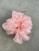 A delicate, fluffy pink scrunchie from Noémiah's "Three Scrunchies" collection, crafted from 100% silk organza, is elegantly positioned on a smooth gray surface. With its airy, ruffled appearance reminiscent of a tulip dress, the scrunchie exudes elegance and grace.