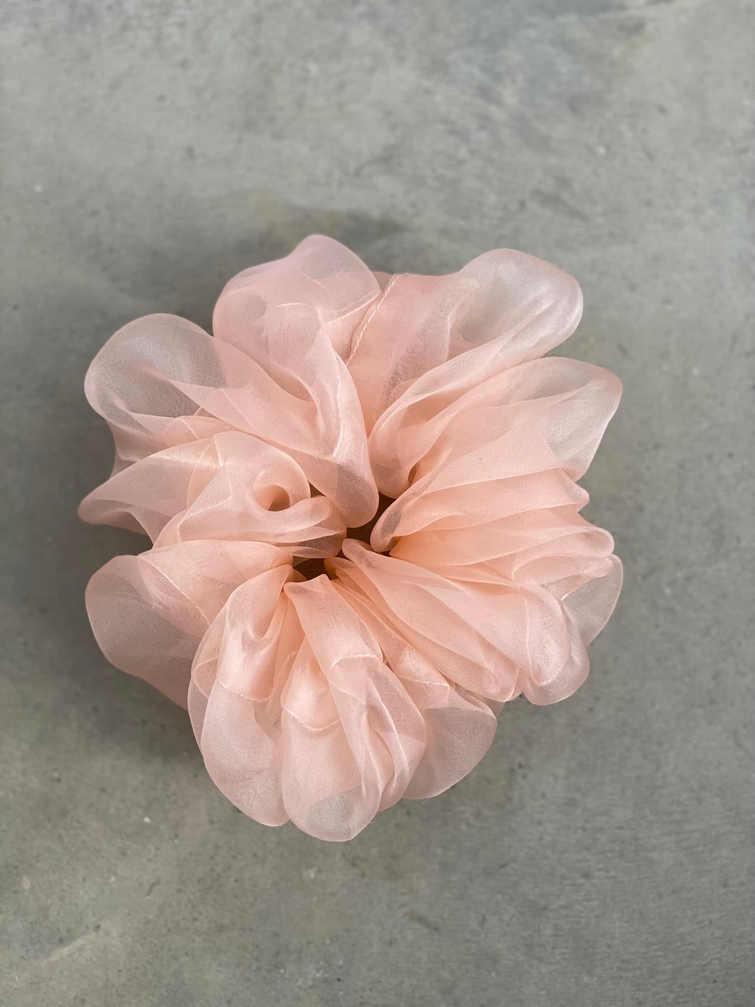 A close-up of a pink, sheer tulle scrunchie from the "Three Scrunchies" collection by Noémiah is displayed on a gray textured background. Crafted from 100% silk organza, this delicate accessory boasts a puffy appearance reminiscent of layers of flowers and resembles Tulip Dress scraps.