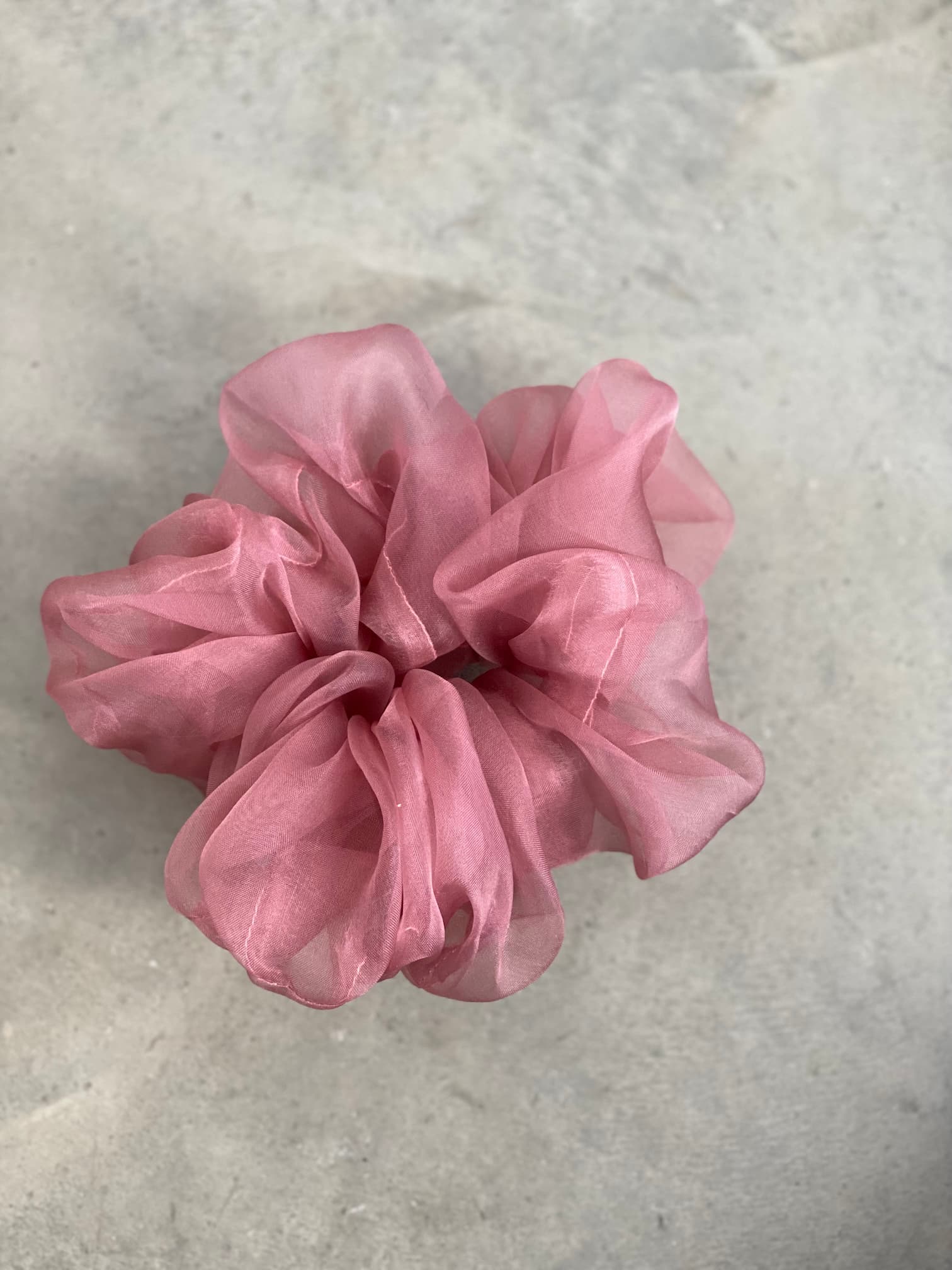 A pink, sheer fabric scrunchie from Noémiah's "Three Scrunchies" collection, made from tulip dress scraps, rests elegantly on a gray textured surface.