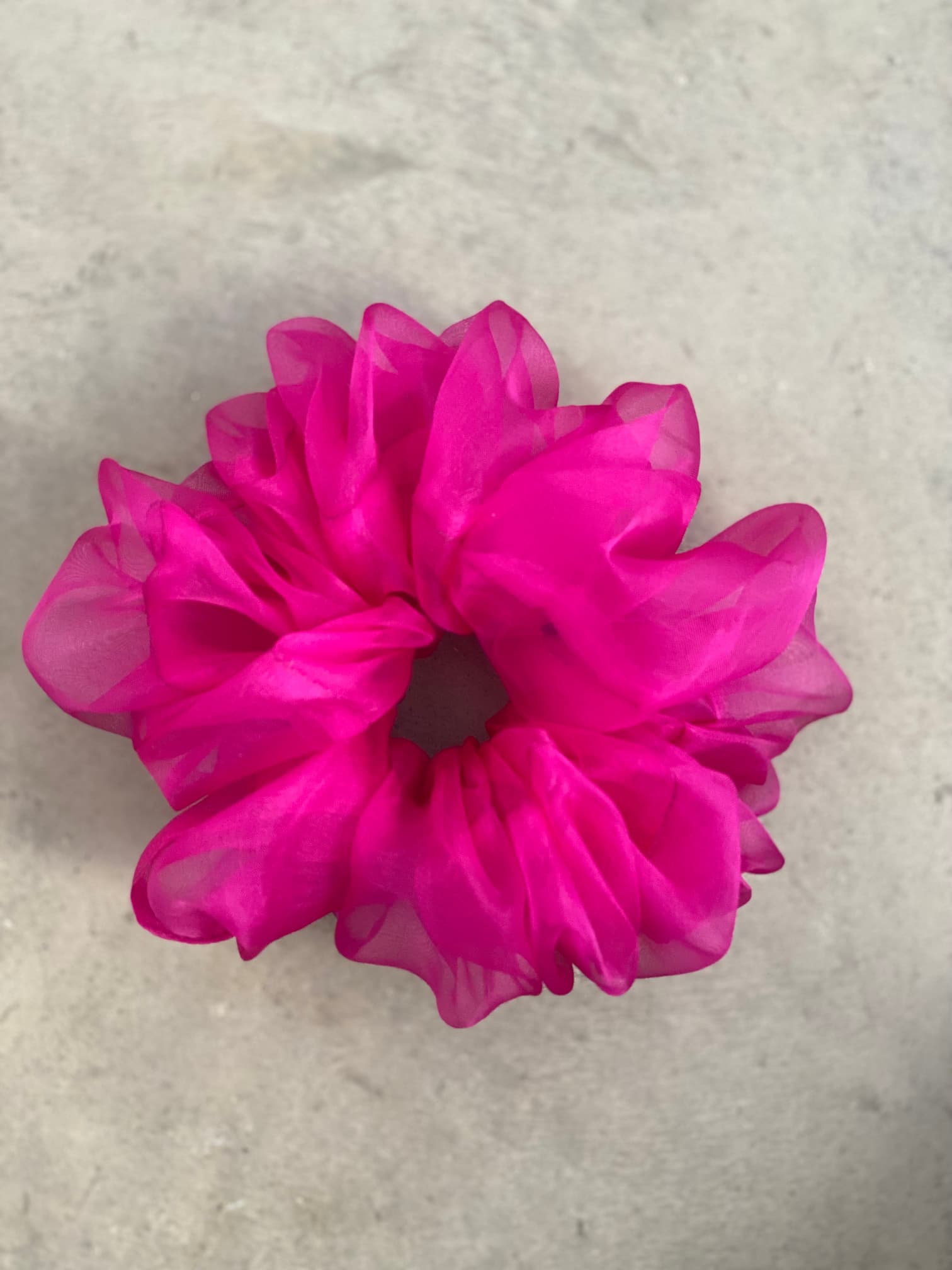 A vibrant pink scrunchie from the "Three Scrunchies" collection by Noémiah, made from 100% silk organza with elegant ruffled layers, is elegantly displayed on a gray textured surface.