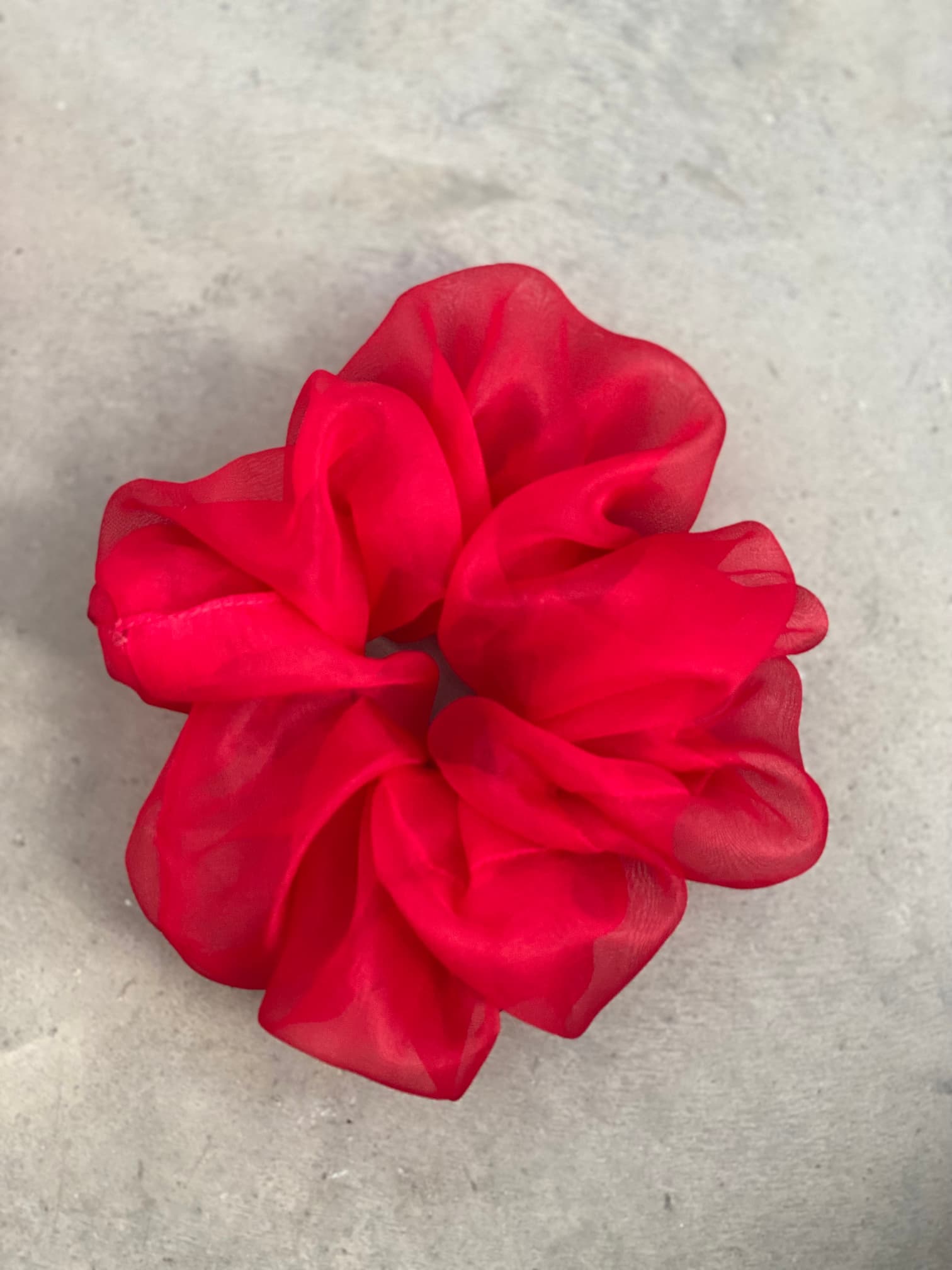 A vibrant red scrunchie from Noémiah's "Three Scrunchies" collection, crafted from 100% silk organza with a sheer and ruffled texture, rests elegantly on a light gray, textured surface.