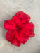 A vibrant red scrunchie from Noémiah's "Three Scrunchies" collection, crafted from 100% silk organza with a sheer and ruffled texture, rests elegantly on a light gray, textured surface.