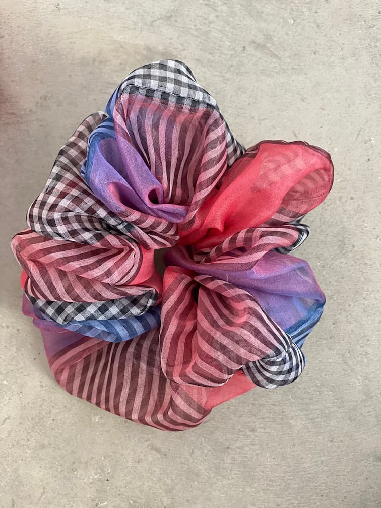 A vibrant, see-through scrunchie made from Tulip Dress remnants by Noémiah blends pink, blue, and checkered designs on a light gray background. This is part of their Three Scrunchies collection.