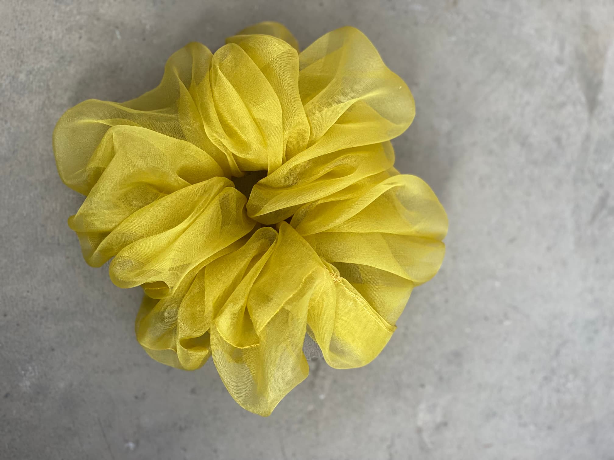 A large, fluffy yellow scrunchie from the "Three Scrunchies" set by Noémiah is placed on a concrete surface. Carefully crafted from 100% silk organza, the sheer and layered fabric gives it a voluminous appearance.