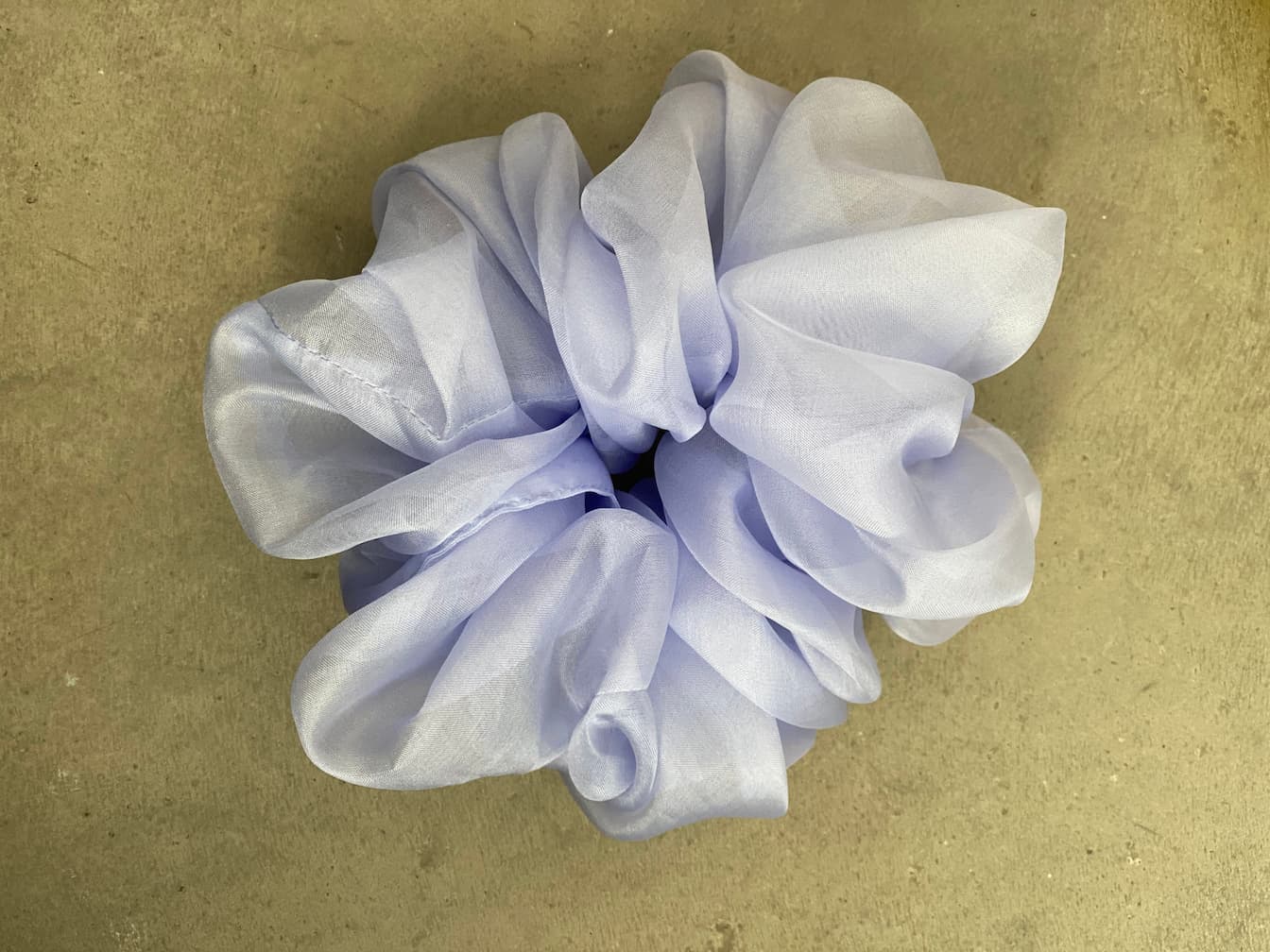 The light purple scrunchie from Noémiah's "Three Scrunchies" collection, made entirely of silk organza, showcases a voluminous and ruffled design against a simple, light-hued backdrop.