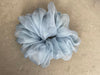 A large, fluffy scrunchie from Noémiah's Three Scrunchies collection sits gracefully on the concrete surface. Crafted from 100% silk organza, it boasts a light blue, translucent appearance in a loose, circular shape with gentle folds and ruffles reminiscent of delicate Tulip Dress scraps.