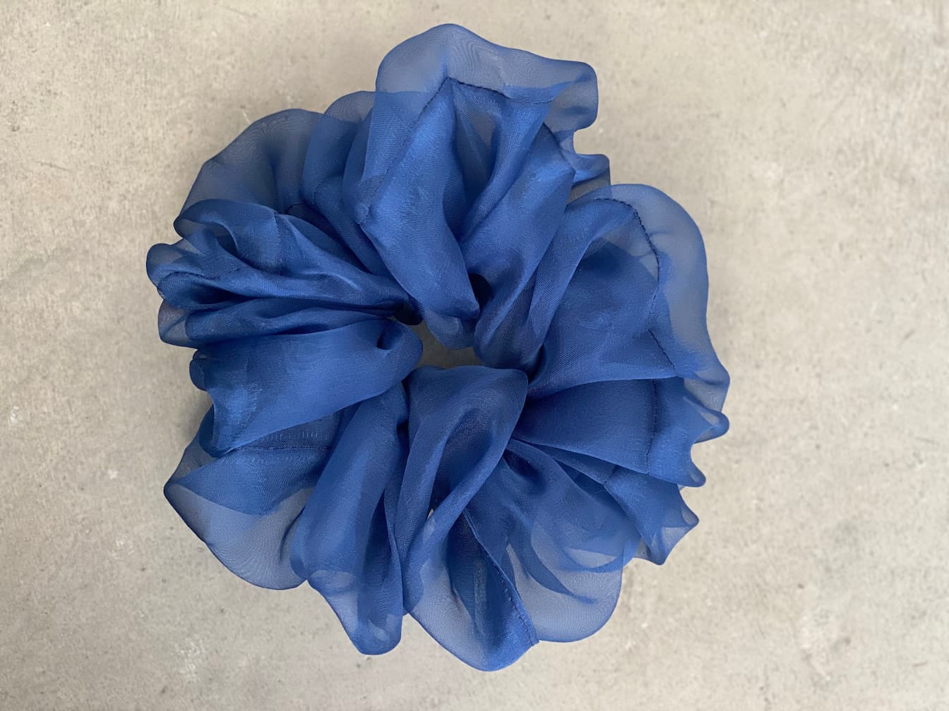 A large blue scrunchie, part of the "Three Scrunchies" set by Noémiah, crafted from Tulip Dress scraps, is displayed on a light gray surface. The chiffon fabric is gathered in soft, flowing folds, giving it a voluminous and ruffled appearance.