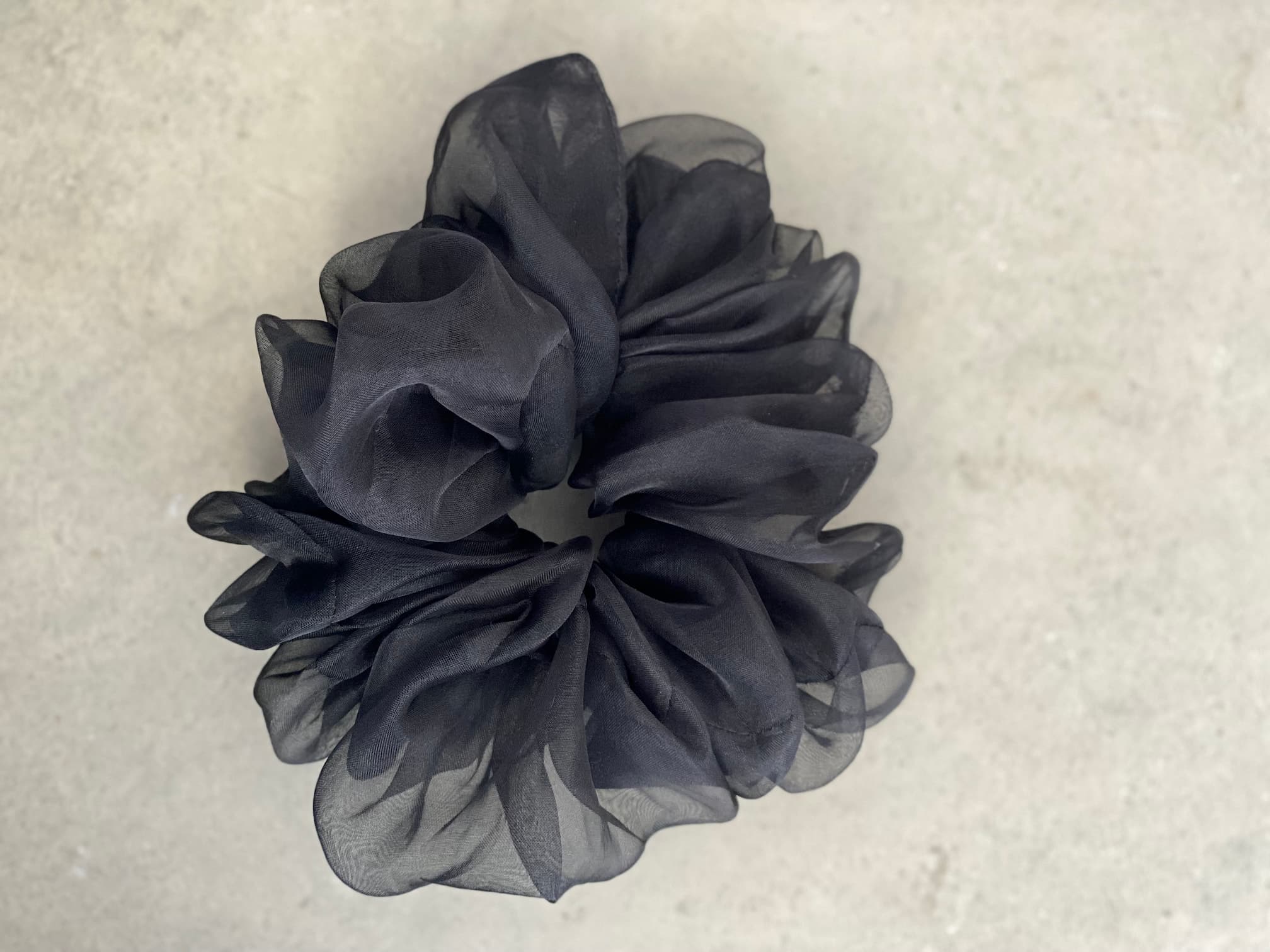 A voluminous, black silk organza scrunchie with a ruffled texture from the "Three Scrunchies" collection by Noémiah is set against a light gray, textured background.