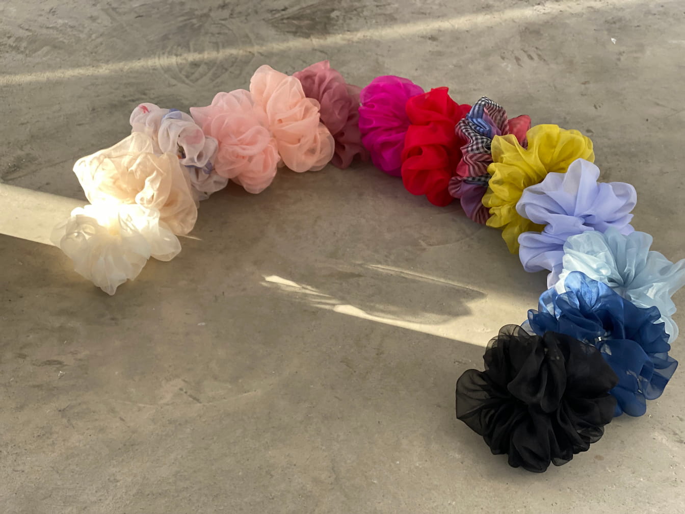 A curved row of vibrant scrunchies is displayed on a concrete surface, showcasing the "Three Scrunchies" by Noémiah. Made from 100% silk organza, these exquisite scrunchies are available in pink, red, yellow, blue, black, and an array of patterned designs. They cast gentle shadows in the sunlight and are crafted uniquely using Tulip Dress scraps.