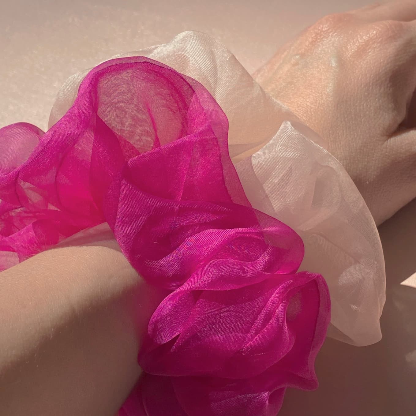 A close-up of a wrist adorned with two sheer, ruffled scrunchies from Noémiah's Three Scrunchies collection, crafted from 100% silk organza. One scrunchie is vibrant pink while the other is pale pink, both made from Tulip Dress scraps. The softly lit background accentuates the fabric's delicate texture.