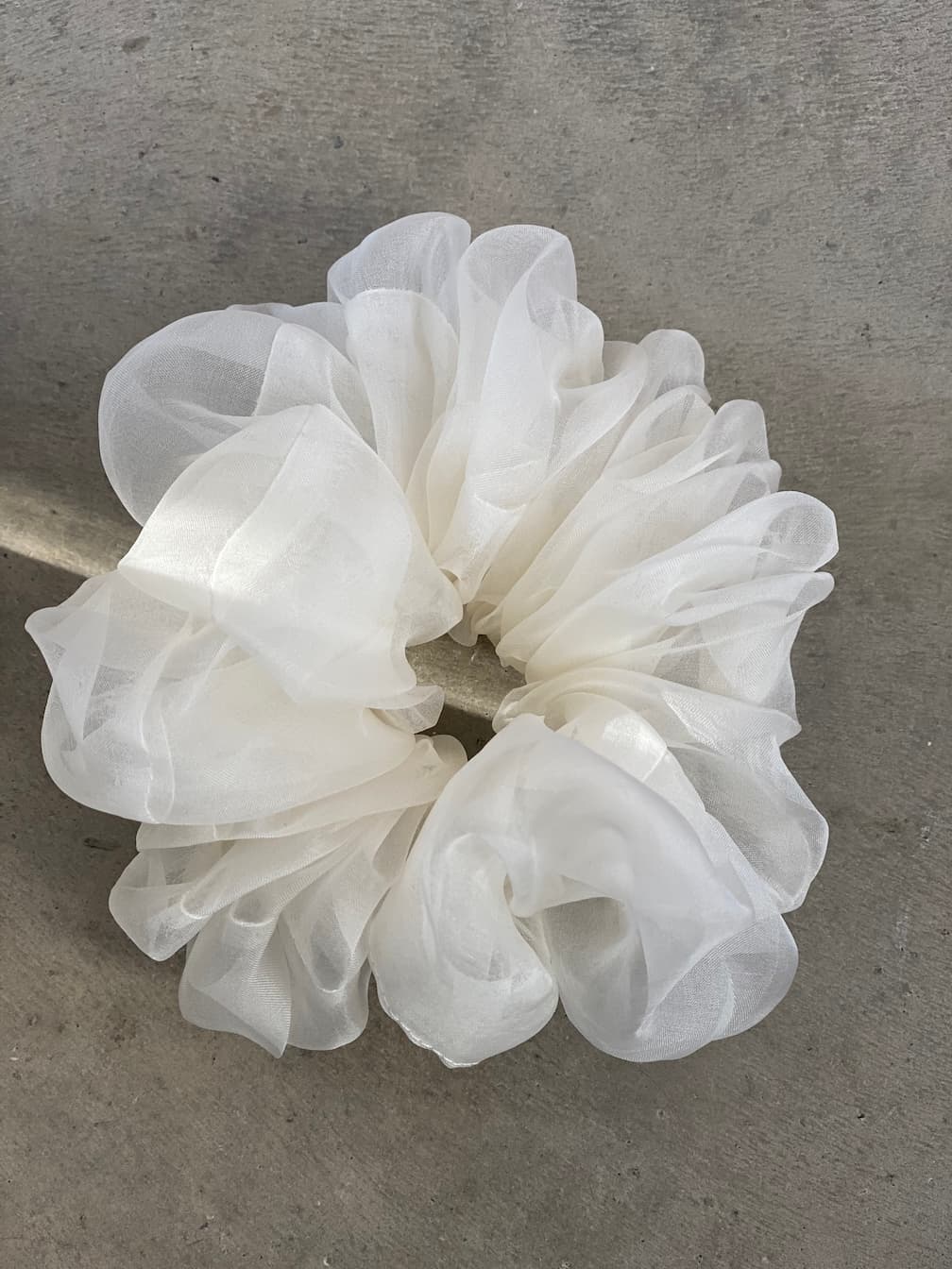 A sizable, fluffy white scrunchie, crafted from sheer and lightweight fabric repurposed from Tulip Dress scraps by Noémiah, rests elegantly on a gray, textured surface.