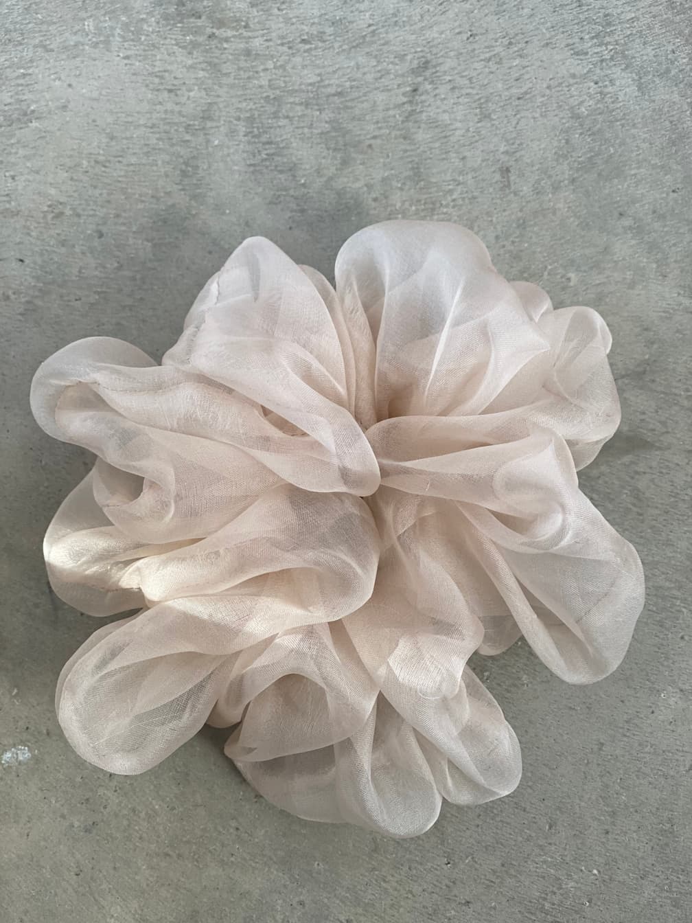 On a smooth, gray surface rests a collection called "Three Scrunchies" by the brand Noémiah. These delicate accessories, crafted from 100% silk organza, display layers gathered to create fluffy, cloud-like formations that whisper luxury and charm in light pink hues.