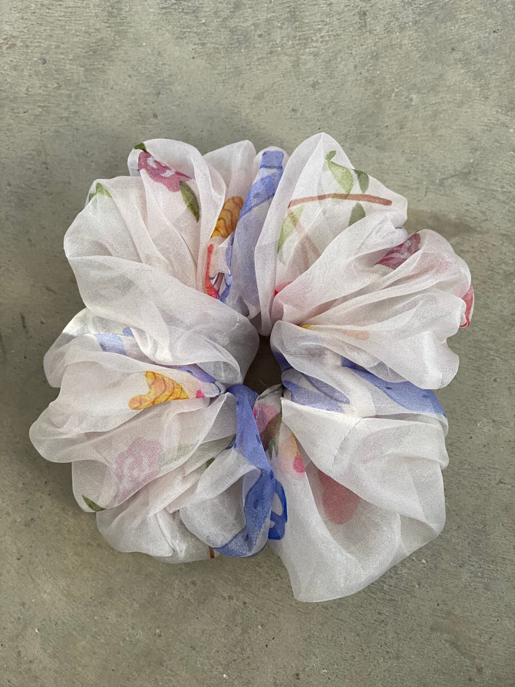 A sheer silk scrunchie from Noémiah's "Three Scrunchies" collection, crafted from 100% silk organza and featuring a floral pattern, rests on a gray surface. The pastel hues—shades of pink, blue, and green—lend it a soft, delicate look reminiscent of Tulip Dress scraps.