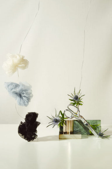 A still life image features "Three Scrunchies" by Noémiah, with tulle puffs elegantly hanging in white, gray, and black. Below them, a block of ice encases scissors and thistle flowers while water gently spills onto a white surface. The scene delicately hints at the texture of 100% silk organza.