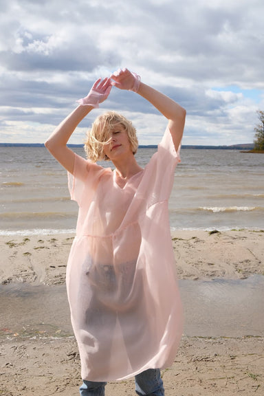 A person stands on the beach in a sheer noemiah Tulip Dress, arms raised as the wind tousles their hair. The sky is partly cloudy, creating a serene and dynamic scene by the water, with the relaxed fit of the silk organza fabric flowing gracefully around them.