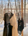 In front of a sunlit window, three sheer garments hang on wooden hangers, featuring a black dress, the Tulip dress by noemiah—a white one-shoulder creation crafted from silk organza—and a black bodysuit adorned with crescent moon designs. Snow and bare trees can be seen outside.