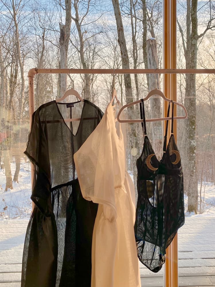 In front of a sunlit window, three sheer garments hang on wooden hangers, featuring a black dress, the Tulip dress by noemiah—a white one-shoulder creation crafted from silk organza—and a black bodysuit adorned with crescent moon designs. Snow and bare trees can be seen outside.