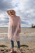 A person dressed in a Tulip Dress by noemiah, crafted from flowing pink silk organza and paired with jeans, stands on a beach with one arm lifted overhead. The partly cloudy sky and ocean serve as the picturesque backdrop.