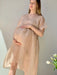 A pregnant woman, dressed in the Tulip Dress by noemiah crafted from sheer silk organza, gently cradles her belly with both hands. She stands bathed in soft natural light against a plain wall, as white tulips can be seen in the lower right corner.