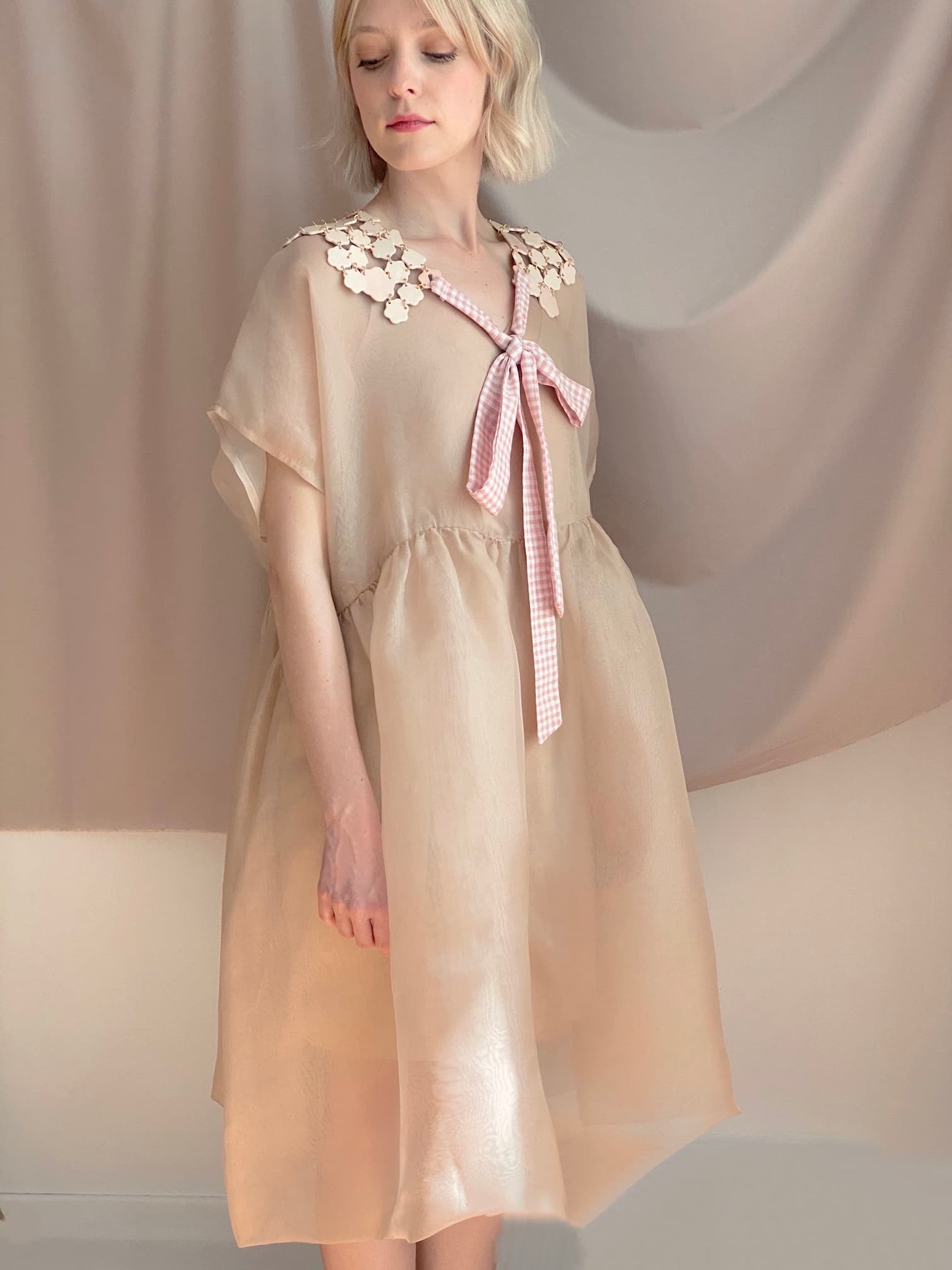 A person with short, light-colored hair stands against a draped fabric background, wearing a sheer Tulip Dress from noemiah in beige, featuring a floral collar and a pink gingham bow. The dress offers a relaxed fit.