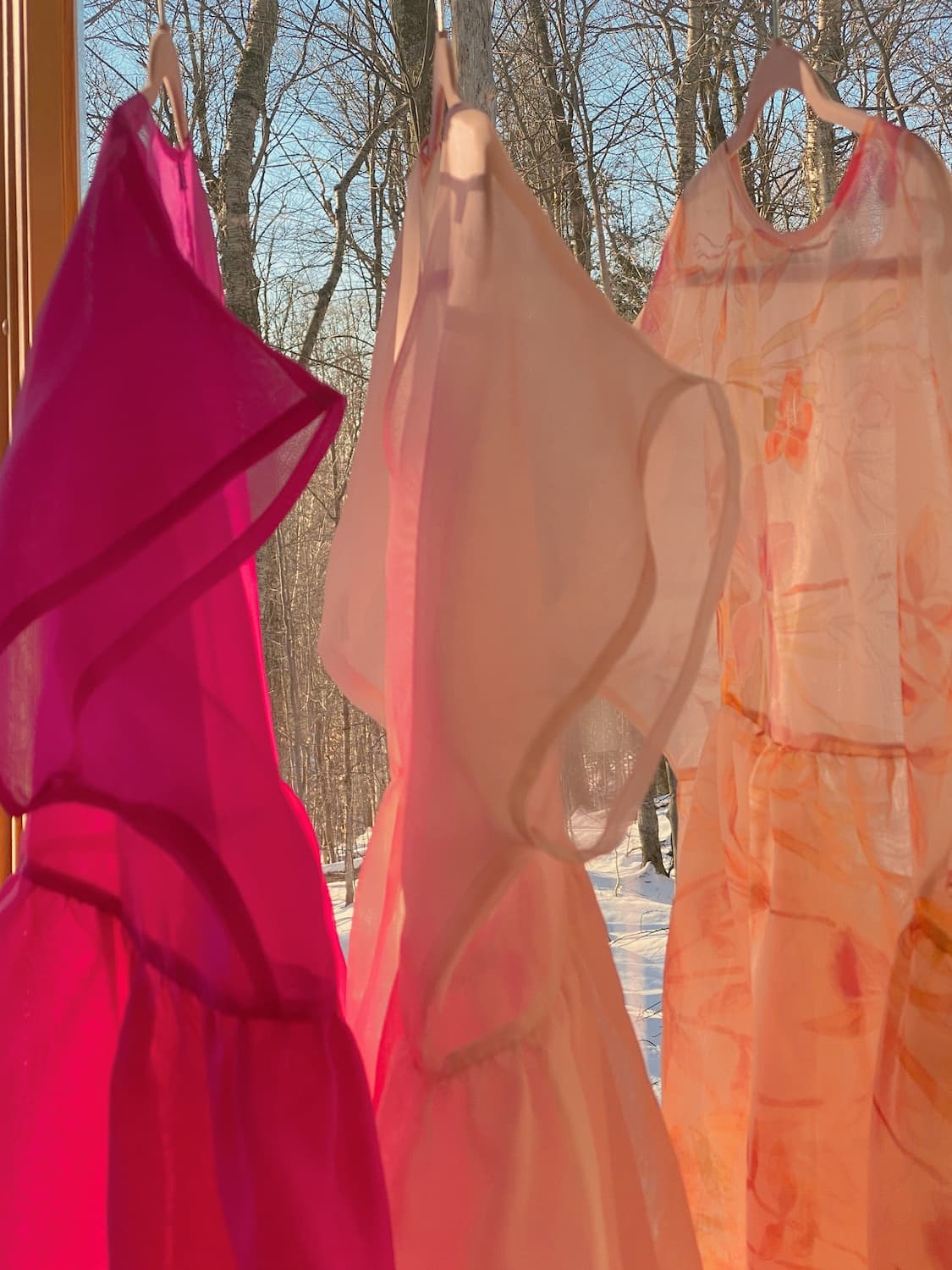 Three vibrant, translucent silk organza dresses from noemiah, featuring shades of pink, peach, and pale yellow, are elegantly displayed on hangers by a window. Sunlight streams through the relaxed-fit fabric, casting a warm glow against the snow-covered trees and clear blue sky. The Tulip Dress from noemiah gracefully stands out amidst this serene scene.