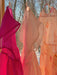 Three vibrant, translucent silk organza dresses from noemiah, featuring shades of pink, peach, and pale yellow, are elegantly displayed on hangers by a window. Sunlight streams through the relaxed-fit fabric, casting a warm glow against the snow-covered trees and clear blue sky. The Tulip Dress from noemiah gracefully stands out amidst this serene scene.