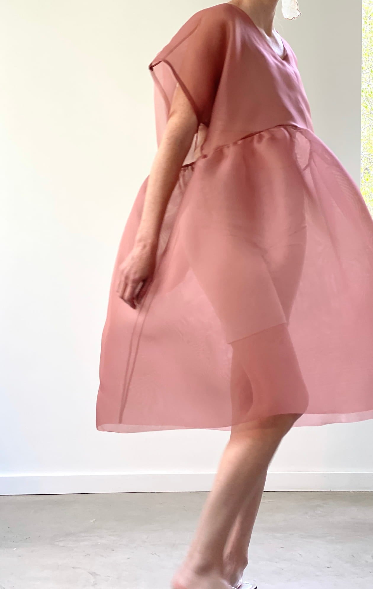 A person wearing a flowing sheer pink Tulip Dress by noemiah walks across a brightly lit room. The translucent silk organza fabric offers a relaxed fit, beautifully showcasing movement and lightness. The minimal, soft background enhances the delicate nature of the outfit.