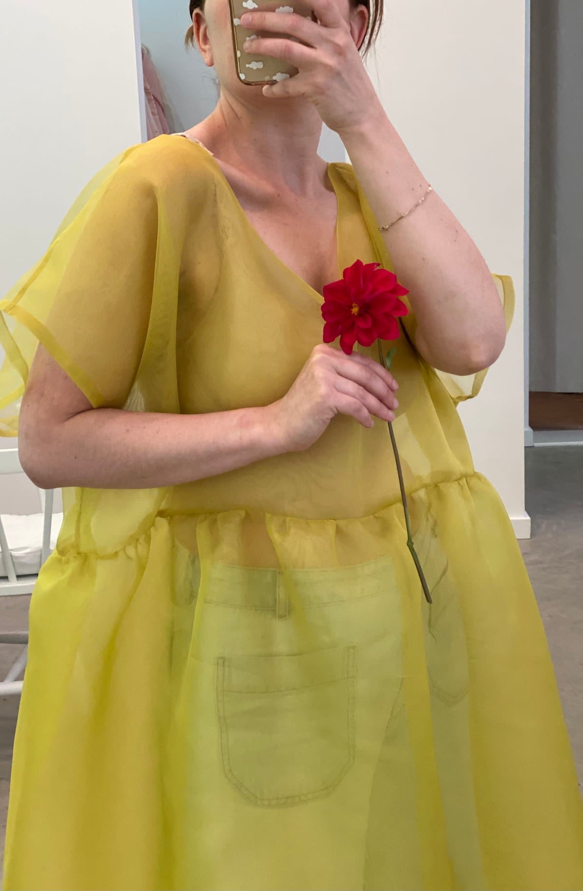 A person takes a mirror selfie wearing the sheer yellow Tulip Dress by noemiah, holding a red flower with their face partially hidden by a phone. The silk organza dress, showcasing visible pockets and a relaxed fit, stands out against the minimalistic background.