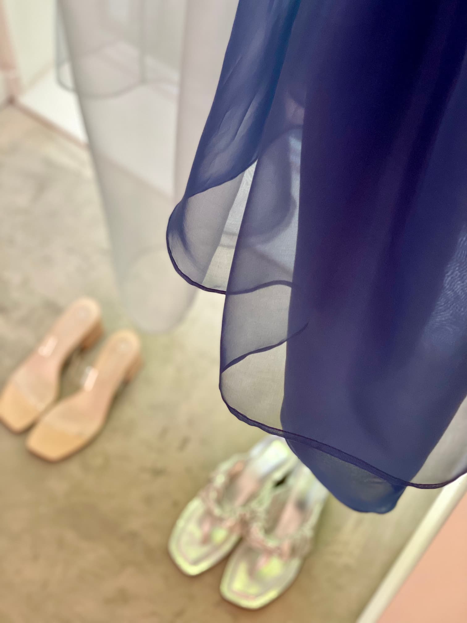 A close-up of the noemiah Tulip Dress, crafted from delicate silk organza in a sheer blue hue, hangs gracefully above two pairs of shoes on a tan floor. One pair features beige, open-toed block heels, while the other showcases silver with shiny, intricate straps. The dress's relaxed fit enhances its ethereal feel.