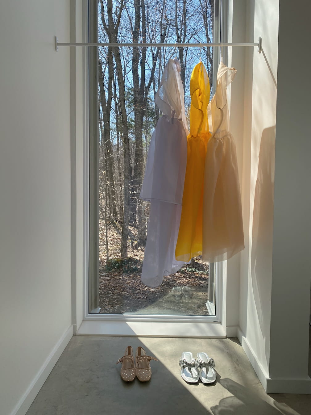 A large window offers a view of leafless trees. In front, a relaxed fit white dress and a silk organza yellow Tulip Dress from noemiah hang on a rack. Below them, a pair of beige shoes and silver shoes are placed on the concrete floor.