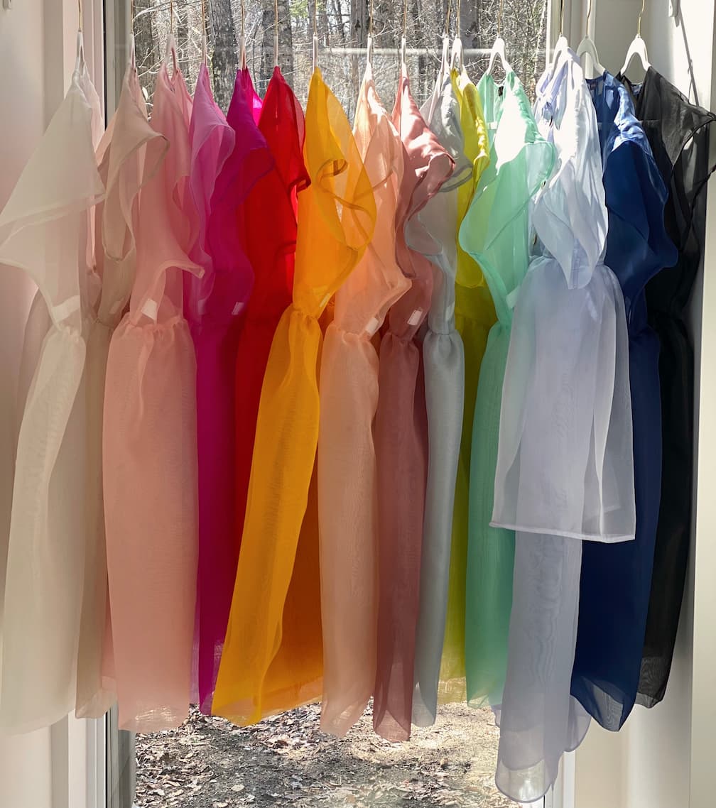 A row of vibrant, semi-transparent silk organza dresses from noemiah hangs on white hangers in front of a window. These Tulip Dresses come in an array of colors including pink, red, orange, yellow, green, blue, and purple. Their relaxed fit is beautifully highlighted by the bright natural light.