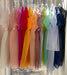 A row of vibrant, semi-transparent silk organza dresses from noemiah hangs on white hangers in front of a window. These Tulip Dresses come in an array of colors including pink, red, orange, yellow, green, blue, and purple. Their relaxed fit is beautifully highlighted by the bright natural light.