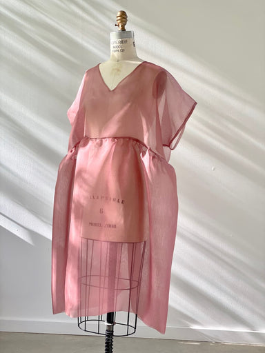 A delicate Tulip Dress by noemiah, expertly crafted from pink sheer silk organza, is showcased on a dress form, its soft silhouette casting a gentle shadow on the white wall. The design includes a relaxed V-neckline and loose, flowing fabric that exudes an airy elegance.