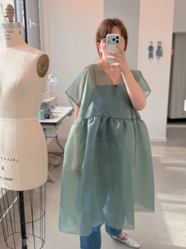 Indoors, a person takes a mirror selfie wearing the noemiah Tulip Dress—a sheer, loose green silk organza style—over jeans with silver shoes. The feminine silhouette stands out, with a dress form and sewing machine behind them.
