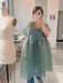 Indoors, a person takes a mirror selfie wearing the noemiah Tulip Dress—a sheer, loose green silk organza style—over jeans with silver shoes. The feminine silhouette stands out, with a dress form and sewing machine behind them.