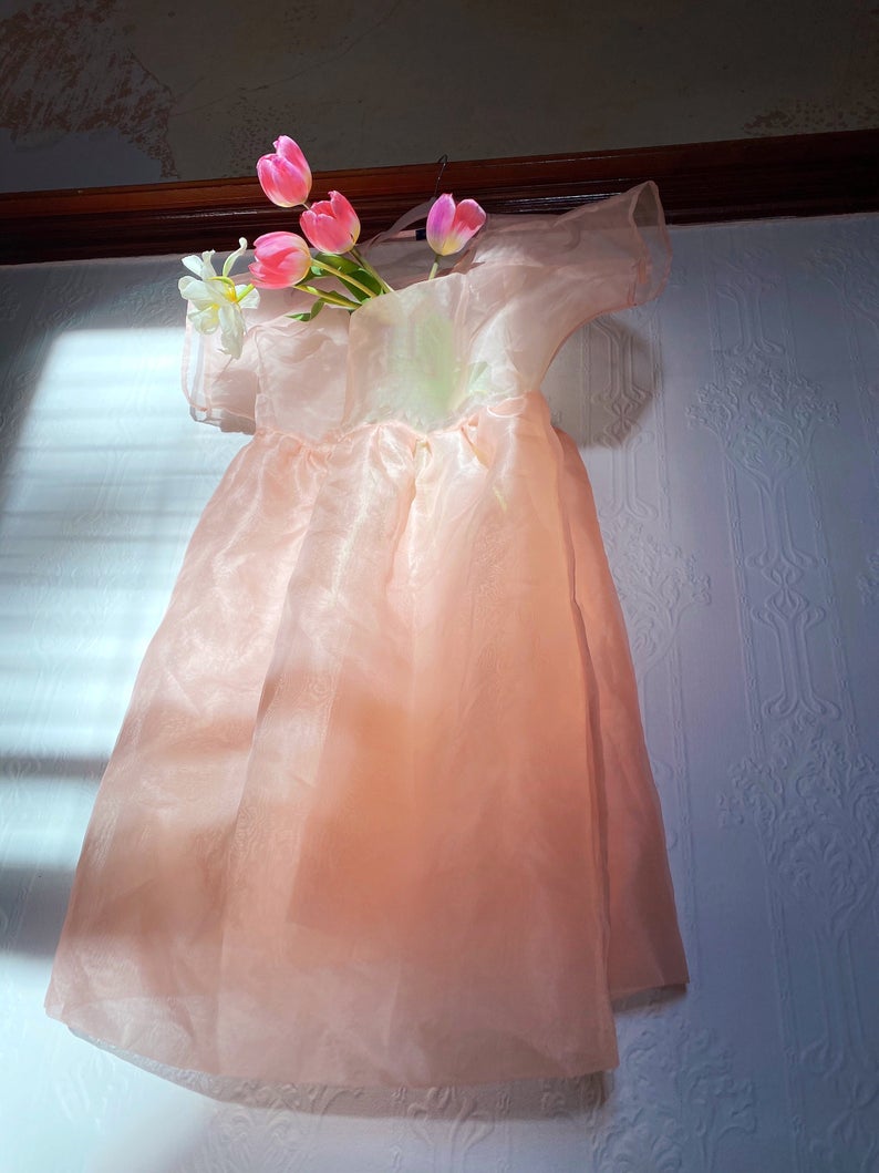 A delicate noemiah Tulip Dress crafted from silk organza is displayed against a textured white wall, bathed in soft sunlight filtering through a nearby window. The dress features three pink tulips thoughtfully arranged in the bodice, lending a whimsical floral accent to its relaxed fit.