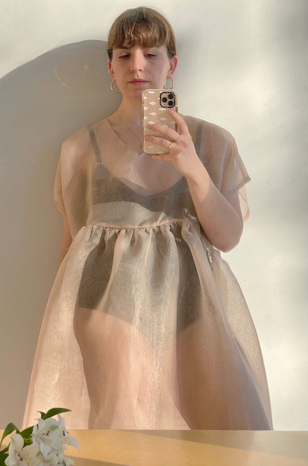 Taking a mirror selfie indoors, this person wears a sheer, beige Tulip Dress from noemiah over a dark top. They hold a phone with a patterned case, while soft sunlight casts shadows on the wall. A small bouquet of white flowers graces the nearby surface, adding charm to this serene moment.