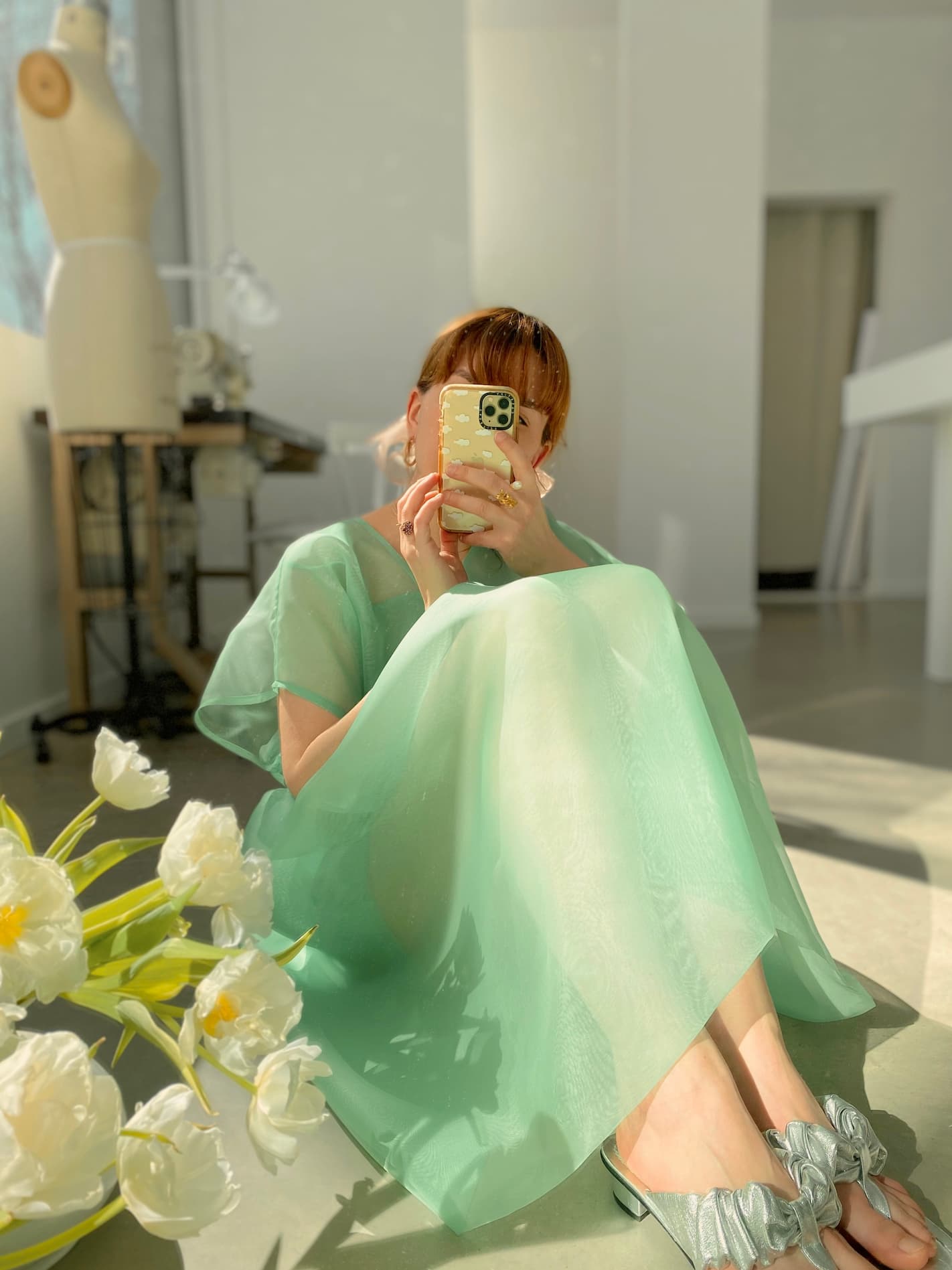 A person wearing the noemiah Tulip Dress, a relaxed-fit sheer piece in light green, sits on the floor capturing a mirror selfie. White tulips adorn the foreground while a dress form in the bright room imparts an elegance reminiscent of silk organza.