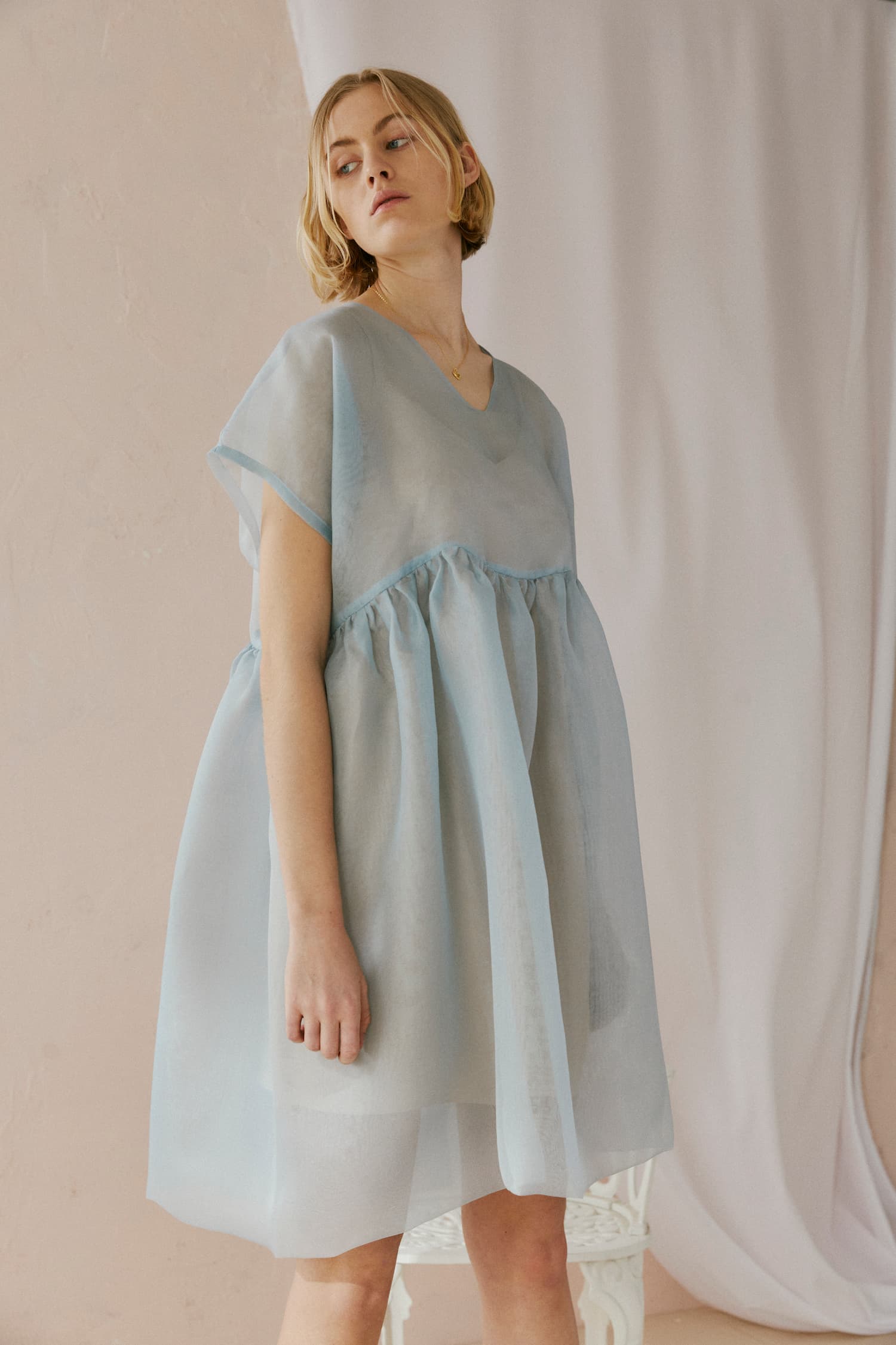 A person stands wearing a loose-fitting, light blue Tulip Dress by noemiah, gazing slightly to the side. The sheer and flowy dress features silk organza that adds elegance to its gathered waist. The background is a neutral space with soft drapery.