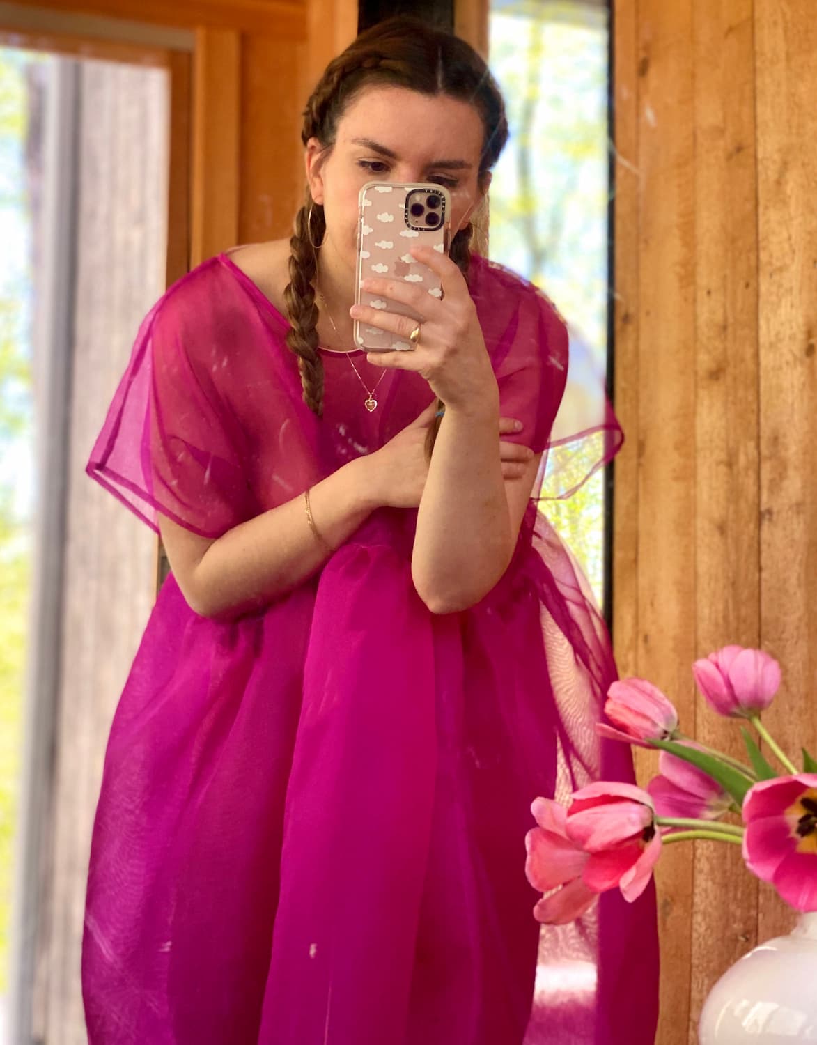 A person wearing the Tulip Dress from noemiah, a bright pink sheer silk organza creation, takes a mirror selfie. They have braided hair and stand in a wooden room with pink tulips displayed in a white vase on the right side of the image.