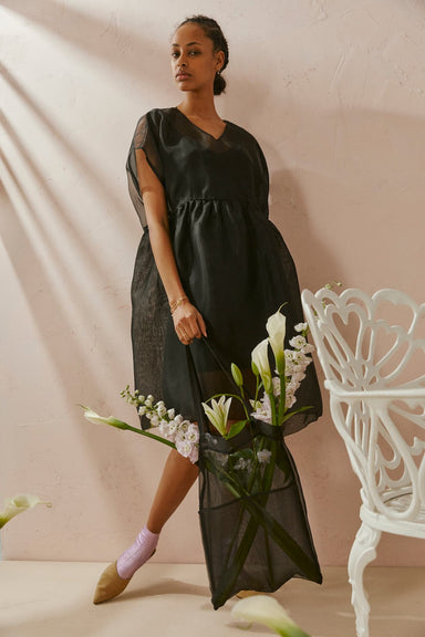 A woman in the noemiah Tulip Dress, a relaxed fit, flowy black dress, leans against a white chair while holding a bouquet of white lilies and purple flowers. She pairs her elegant silk organza attire with tan shoes and purple socks. The minimalist setting, featuring soft lighting and a pale pink wall, highlights her sophisticated style.