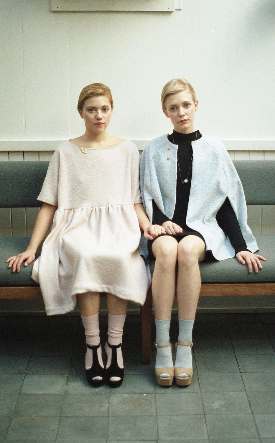 On a minimalist-designed bench, two people with similar hairstyles sit holding hands. One is dressed in a light pink dress paired with light socks and heels, while the other wears a light blue coat over a black dress complemented by chunky shoes. Their harmonious appearance is accentuated by the stylish addition of the Urso Necklace from noemiah.