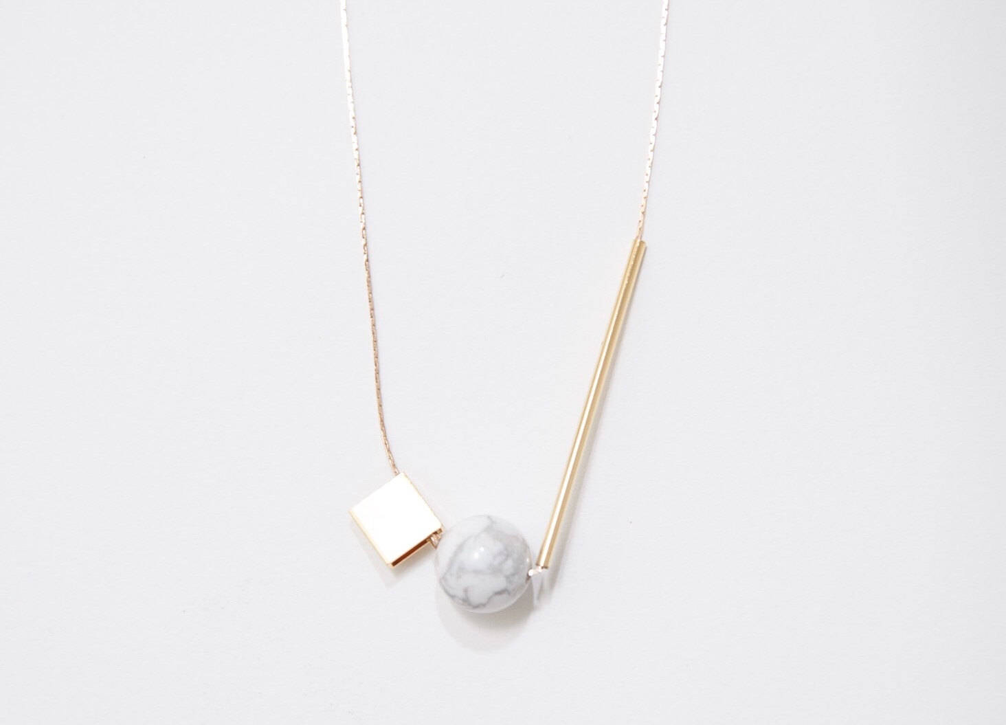 The Urso Necklace by noemiah is a minimalist piece featuring tarnish-resistant plating, a thin gold chain, and a geometric pendant showcasing a gold square, white marble-like sphere, and gold rod.