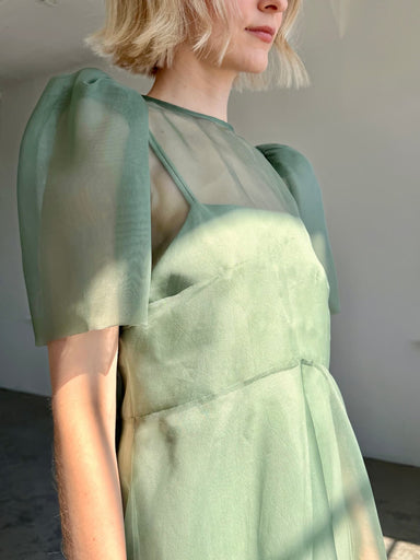 A person with short blonde hair wears the Noémiah Victoire Dress, a sheer silk organza midi dress with puff sleeves, standing in soft sunlight against a neutral background.