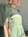 A person with short blonde hair wears the Noémiah Victoire Dress, a sheer silk organza midi dress with puff sleeves, standing in soft sunlight against a neutral background.
