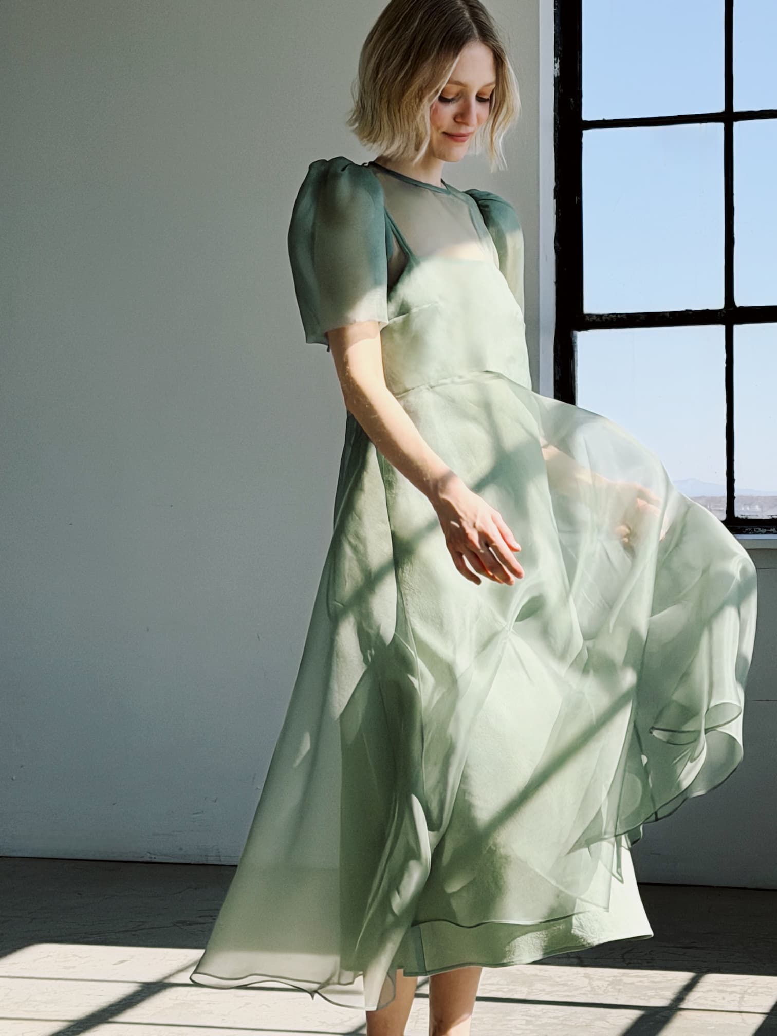 A woman in the Noémiah Victoire Dress—light green, semi-sheer silk organza with puff sleeves—stands by a sunlit window, gently holding the skirt and smiling softly.