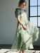 A woman in the Noémiah Victoire Dress—light green, semi-sheer silk organza with puff sleeves—stands by a sunlit window, gently holding the skirt and smiling softly.