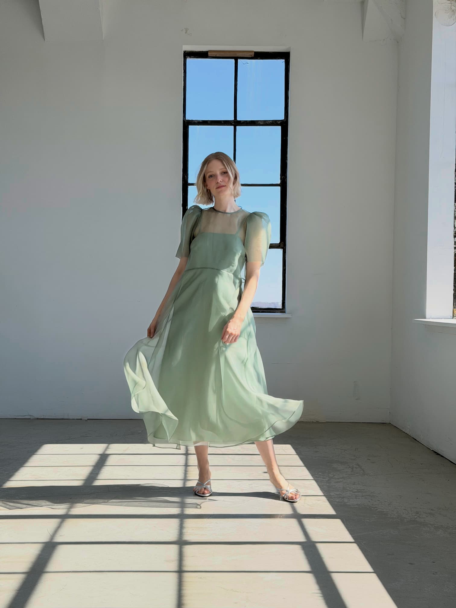 A woman wears the Noémiah Victoire Dress, a light green silk organza midi with puff sleeves, standing gracefully in a sunlit room before a tall window as sunlight casts geometric shadows on the floor.
