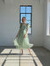 A woman wears the Noémiah Victoire Dress, a light green silk organza midi with puff sleeves, standing gracefully in a sunlit room before a tall window as sunlight casts geometric shadows on the floor.